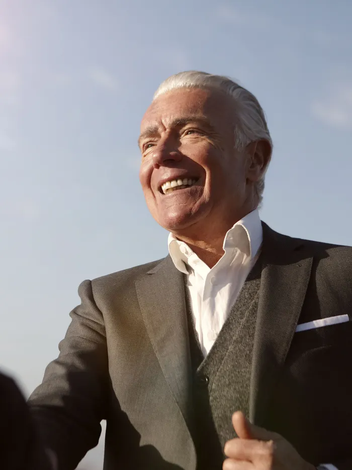 An older man in a suit outdoors, laughing, sky behind him.